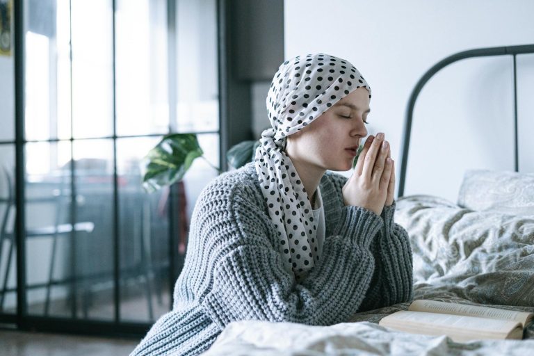 A hopeful woman in a scarf praying in a cozy bedroom during her cancer recovery journey.
