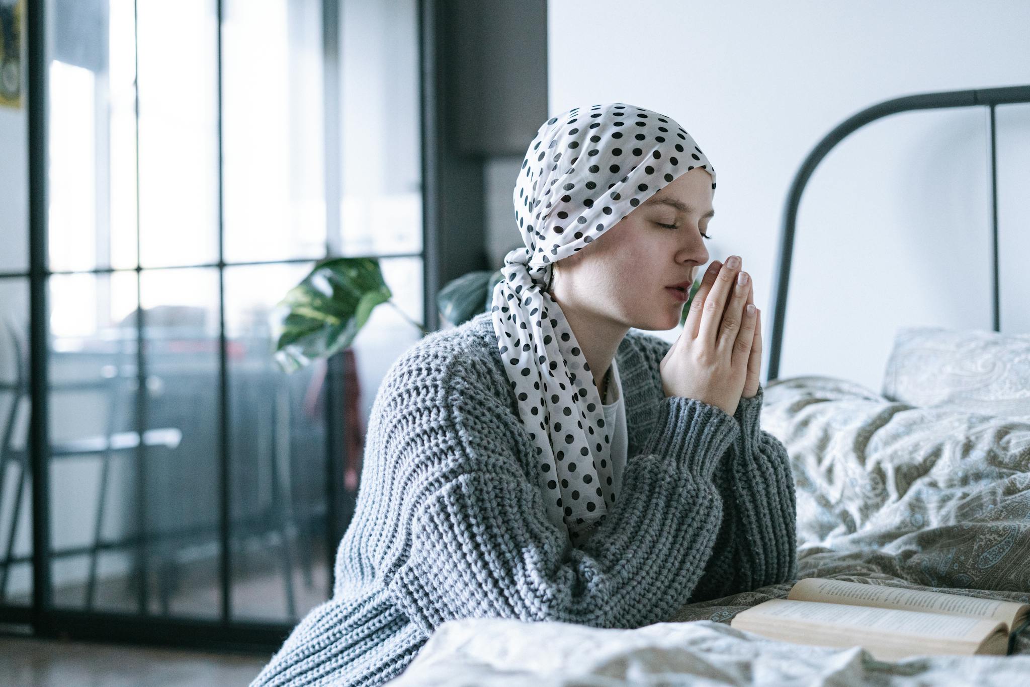 A hopeful woman in a scarf praying in a cozy bedroom during her cancer recovery journey.
